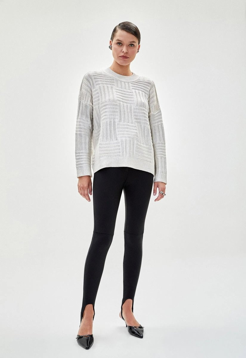 Mulher usando blusa feminina tricot metalizada prata com padrão geométrico e mangas longas. A peça é combinada com legging preta e salto alto.