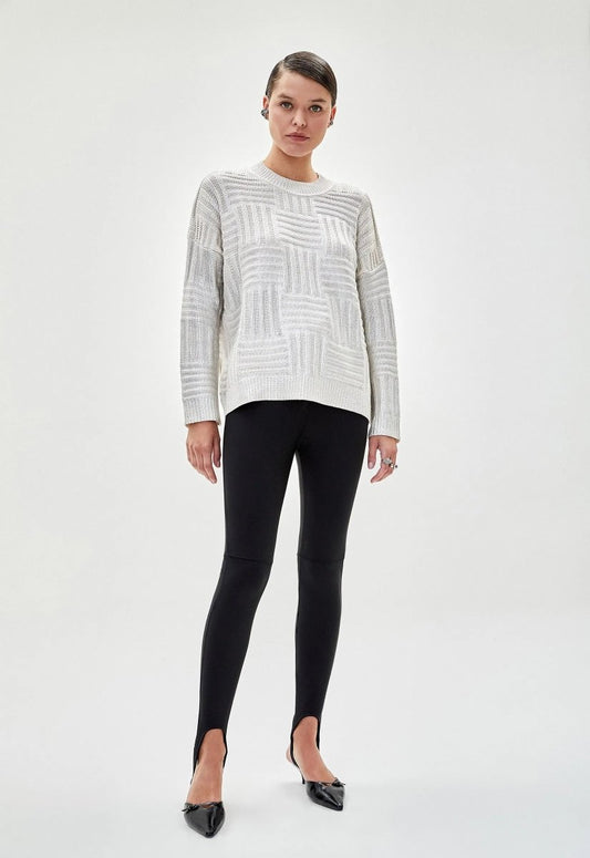 Mulher usando blusa feminina tricot metalizada prata com padrão geométrico e mangas longas. A peça é combinada com legging preta e salto alto.