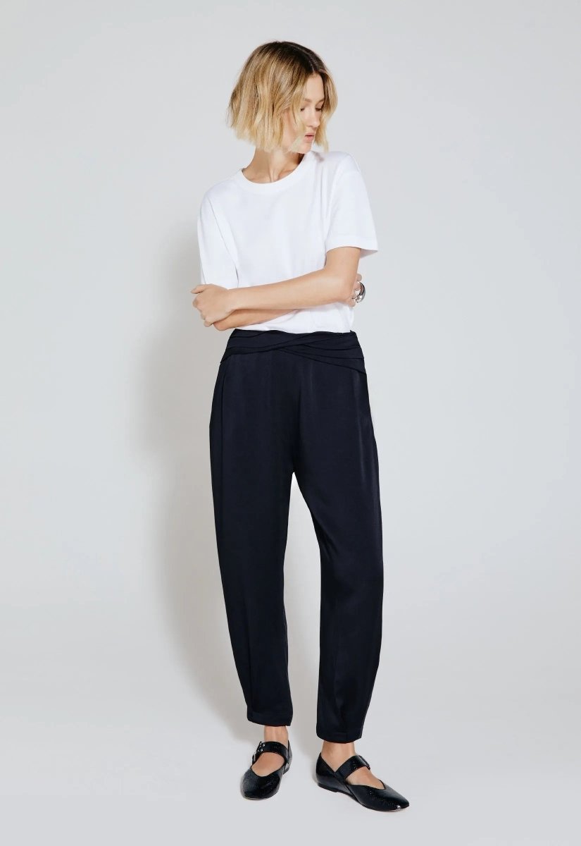 Modelo vestindo calça acetinada preta com caimento fluido, combinada com camiseta branca e sapatos estilo flat pretos.