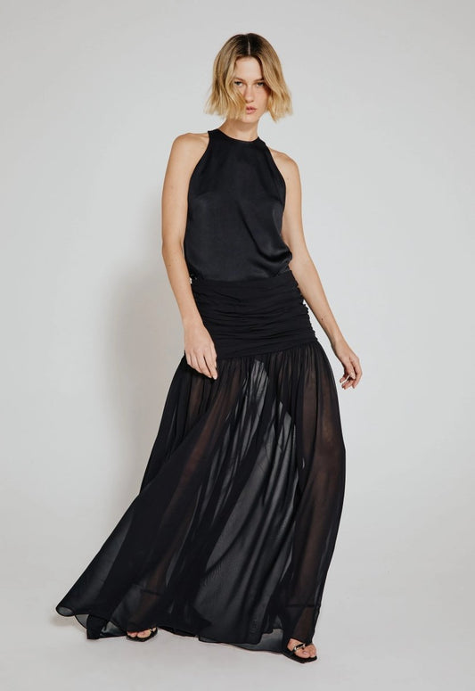 Mulher usando saia longa drapeada preta com transparência, combinada com blusa sem mangas preta. Look moderno e elegante, ideal para ocasiões especiais.