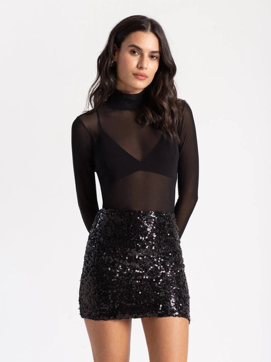 Mulher vestindo saia preta paetê festa com brilho, combinada com blusa de manga longa transparente preta.