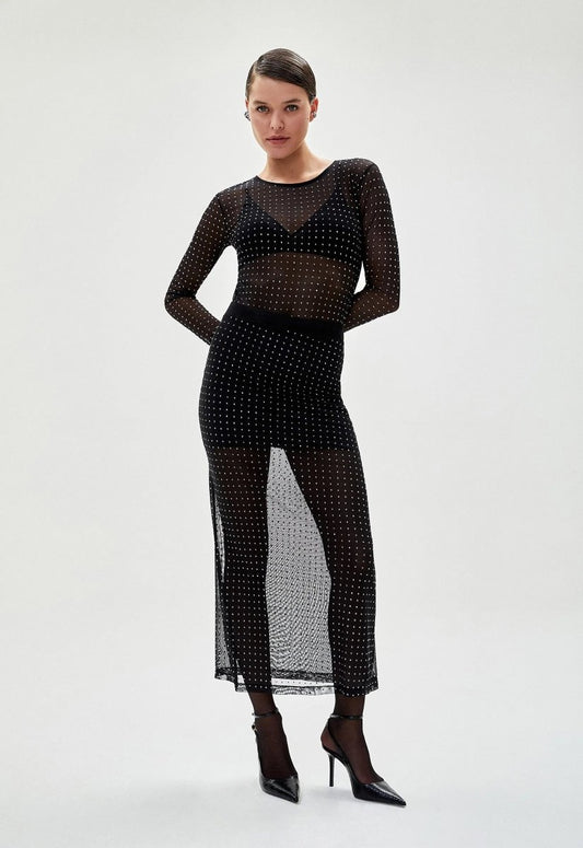 Mulher usando saia tule preta transparente com detalhes brilhantes, combinada com blusa de manga longa no mesmo estilo e salto alto, criando um look sofisticado.