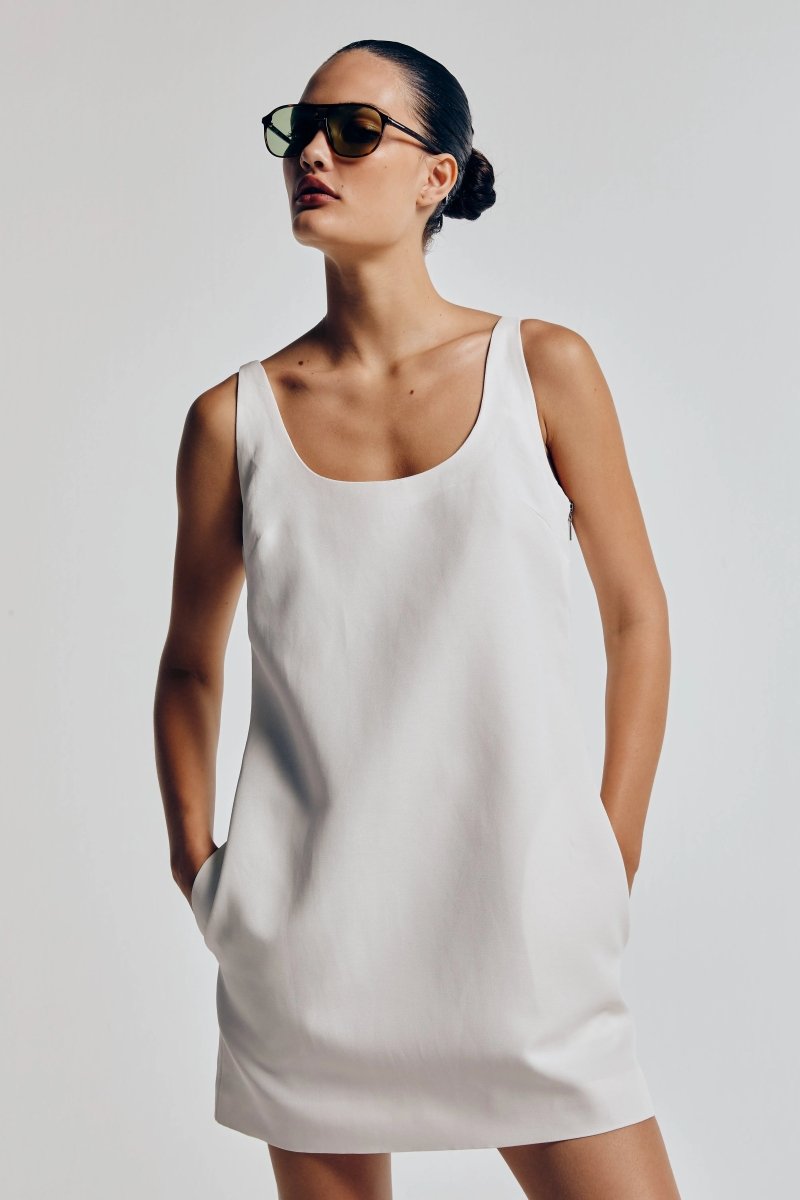 Modelo vestindo vestido linho feminino off white, curto e sem mangas, com decote arredondado. Ela usa óculos escuros e está com cabelo preso.