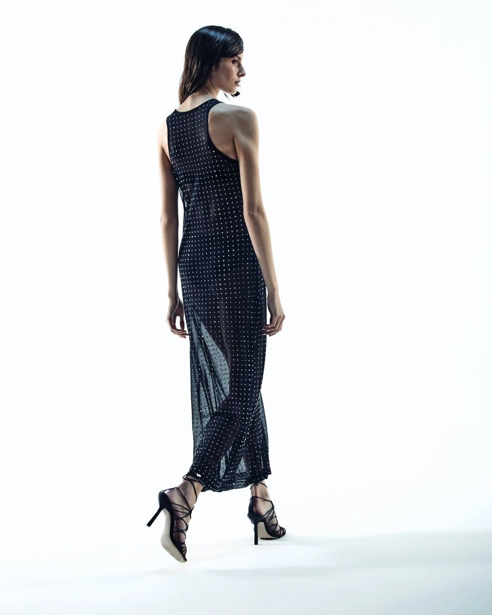 Modelo de costas usando vestido longo preto , com tecido transparente e detalhes brilhantes, combinado com sandálias de salto alto com tiras.