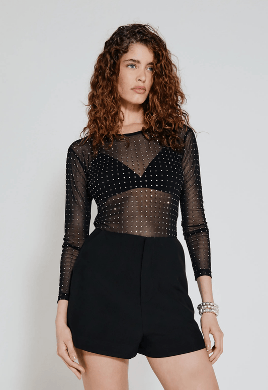 Mulher usando blusa de tule preta com pontos metálicos, shorts pretos ajustados e pulseira prateada.