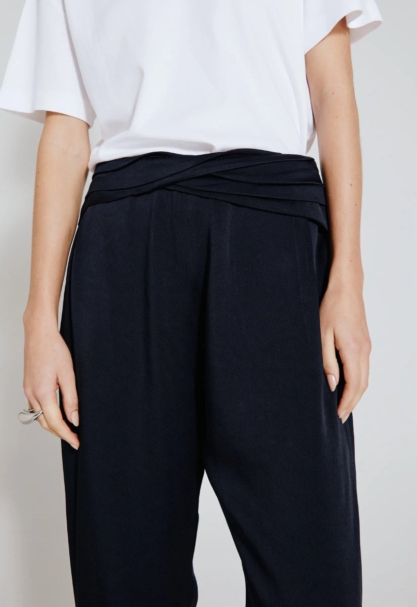 Close de modelo vestindo calça acetinada preta com detalhe transpassado na cintura, combinada com camiseta branca de mangas curtas.