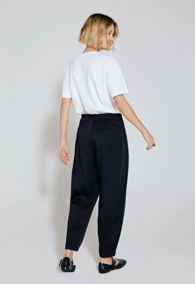 Modelo de costas vestindo calça acetinada preta com cintura alta e detalhe transpassado, combinada com camiseta branca e sapatos pretos estilo flat.