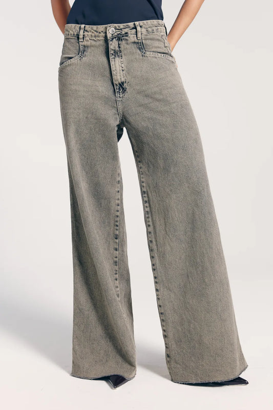 Close da calça jeans feminina reta cintura baixa. A peça possui bolsos e botão dourado. Modelo combina a peça com scarpin preto.