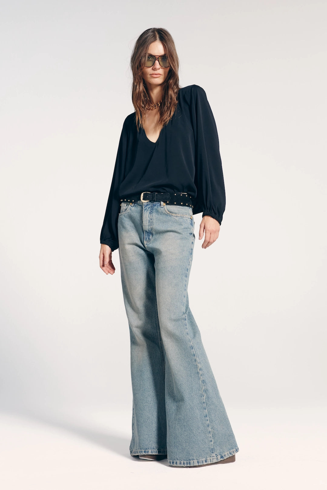 Modelo vestindo calça jeans feminina flare. A peça é combinada com blusa manga longa na cor preta e cinto de fivela preto com tachas douradas.