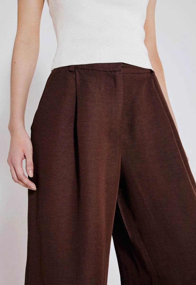 Close da calça marrom wide leg, sofisticada e confortável. Modelo combina a peça com com blusa branca.