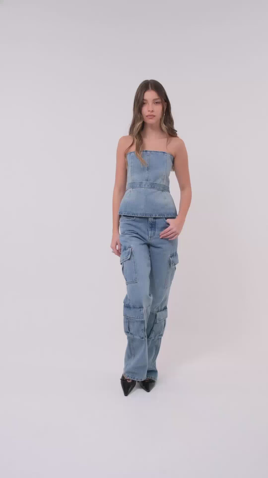 Modelo veste corset jeans com botões dourados na parte de trás. A peça é combinada com calça cargo jeans e scarpin preto.