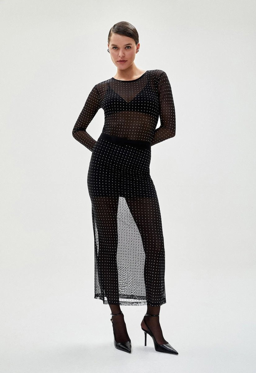 Mulher usando saia tule preta transparente com detalhes brilhantes, combinada com blusa de manga longa no mesmo estilo e salto alto, criando um look sofisticado.