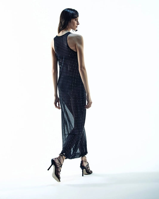 Modelo de costas usando vestido longo  preto , com tecido transparente e detalhes brilhantes, combinado com sandálias de salto alto com tiras.
